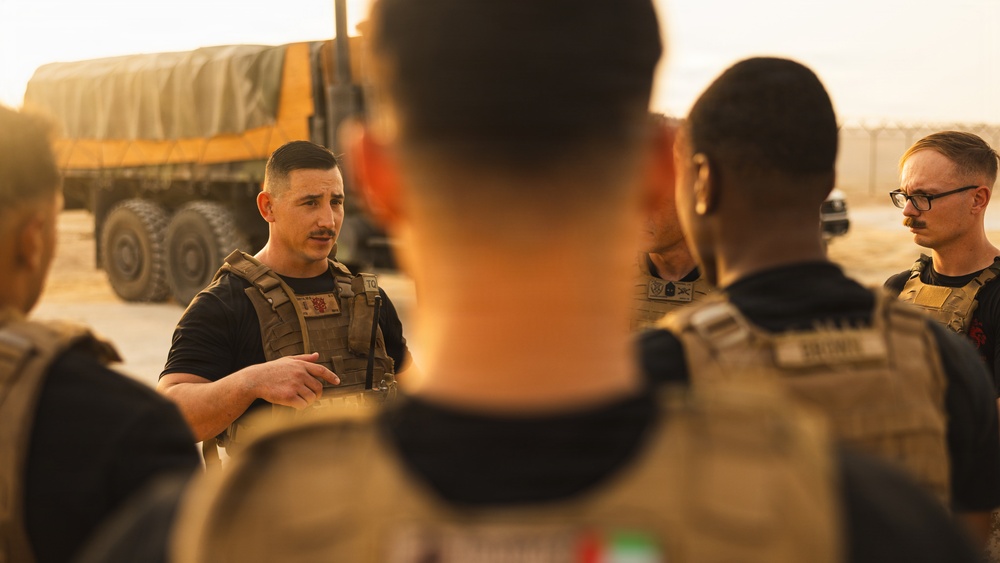 A group of military or security personnel in tactical plate carriers are gathered in a briefing or debriefing formation outdoors. One individual facing the camera appears to be speaking or instructing the others. The setting appears to be an arid, desert-like environment with a large military truck visible in the background.