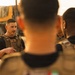 A group of military or security personnel in tactical plate carriers are gathered in a briefing or debriefing formation outdoors. One individual facing the camera appears to be speaking or instructing the others. The setting appears to be an arid, desert-like environment with a large military truck visible in the background.