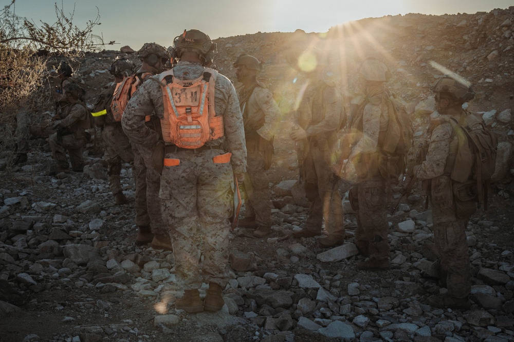 A group of military personnel in desert camouflage uniforms are gathered on rocky terrain during either sunrise or sunset. The central figure is wearing a distinctive orange high-visibility backpack/vest marked with '31M' and the name 'HALL' on their helmet. The scene appears to be a field exercise or operational briefing in an arid environment.