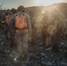 A group of military personnel in desert camouflage uniforms are gathered on rocky terrain during either sunrise or sunset. The central figure is wearing a distinctive orange high-visibility backpack/vest marked with '31M' and the name 'HALL' on their helmet. The scene appears to be a field exercise or operational briefing in an arid environment.
