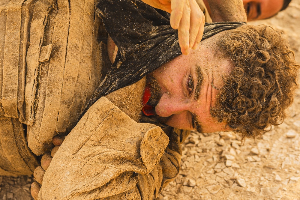 A man with curly hair and a beard is shown lying face-down on rocky, dusty ground, covered in dirt and dust. His face is turned sideways toward the camera, and another person's hand is visible gripping dark fabric near his head. The scene appears to depict a struggle, restraint, or a dramatic/theatrical scenario.