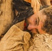 A man with curly hair and a beard is shown lying face-down on rocky, dusty ground, covered in dirt and dust. His face is turned sideways toward the camera, and another person's hand is visible gripping dark fabric near his head. The scene appears to depict a struggle, restraint, or a dramatic/theatrical scenario.
