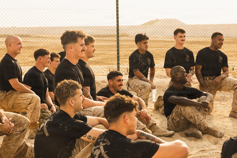 A group of military personnel, appearing to be U.S. Marines based on their distinctive MARPAT camouflage trousers and unit t-shirts, are gathered in a relaxed setting near a chain-link fence in an arid desert environment. The individuals are seated on sandy ground, many smiling and appearing to be in good spirits following what appears to be a physical training or field exercise. Their uniforms show visible dirt and mud, suggesting recent strenuous activity.