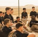 A group of military personnel, appearing to be U.S. Marines based on their distinctive MARPAT camouflage trousers and unit t-shirts, are gathered in a relaxed setting near a chain-link fence in an arid desert environment. The individuals are seated on sandy ground, many smiling and appearing to be in good spirits following what appears to be a physical training or field exercise. Their uniforms show visible dirt and mud, suggesting recent strenuous activity.