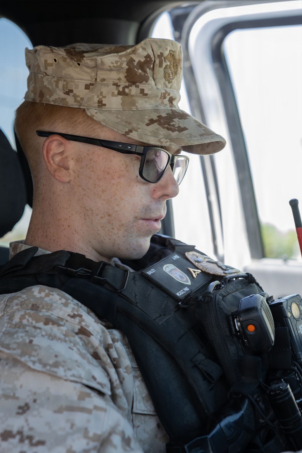 A male military or law enforcement personnel is seated inside a vehicle, wearing desert digital camouflage (MARPAT desert pattern) uniform and cap with a Marine Corps emblem. He is equipped with a tactical vest bearing badges, patches, and what appears to be a body camera with an orange indicator light. He is looking forward, suggesting he may be on patrol or in transit.