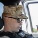 A male military or law enforcement personnel is seated inside a vehicle, wearing desert digital camouflage (MARPAT desert pattern) uniform and cap with a Marine Corps emblem. He is equipped with a tactical vest bearing badges, patches, and what appears to be a body camera with an orange indicator light. He is looking forward, suggesting he may be on patrol or in transit.
