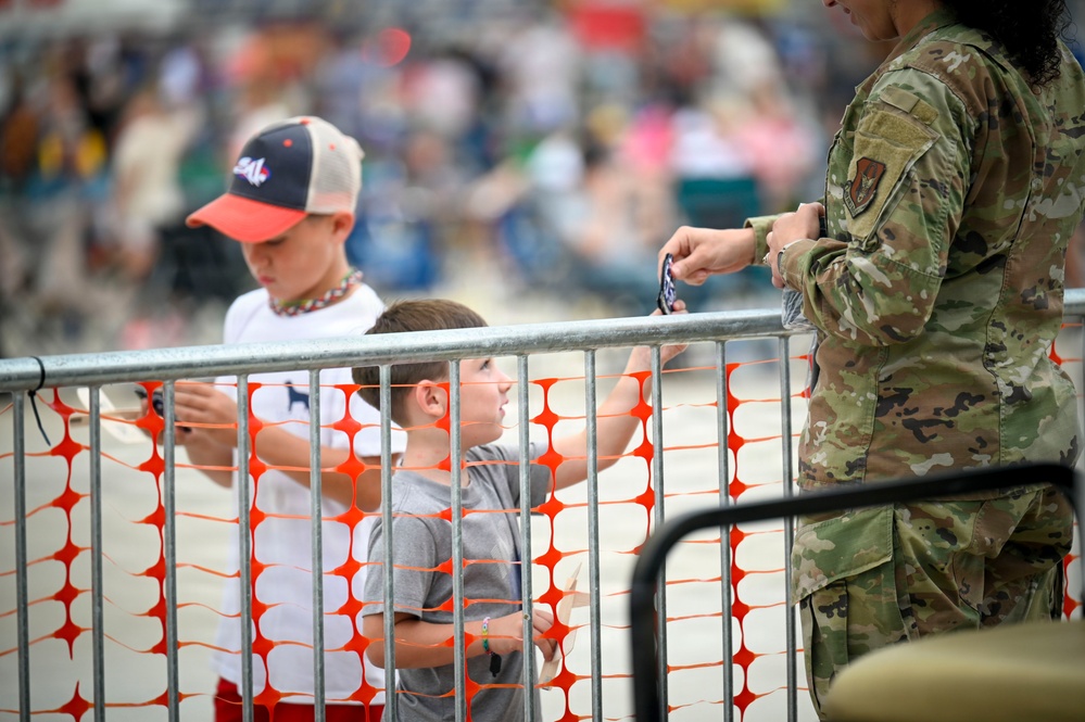 Two young boys stand near a metal crowd-control barrier with orange safety netting at what appears to be an outdoor public event or air show. A military service member in OCP (Operational Camouflage Pattern) uniform stands on the other side of the barrier, appearing to interact with or check something near the children. A large crowd of spectators is visible in the blurred background.