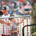 Two young boys stand near a metal crowd-control barrier with orange safety netting at what appears to be an outdoor public event or air show. A military service member in OCP (Operational Camouflage Pattern) uniform stands on the other side of the barrier, appearing to interact with or check something near the children. A large crowd of spectators is visible in the blurred background.