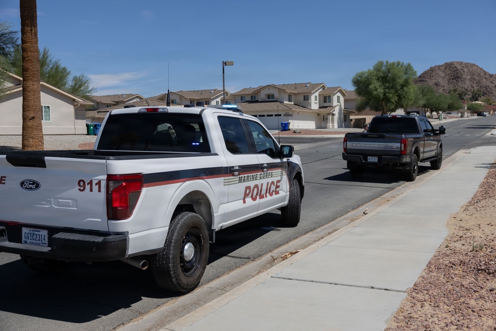 A Marine Corps Police Ford F-150 pickup truck is parked or slowly moving along a residential street in what appears to be a desert Southwest location, likely a military base housing area. The vehicle is marked with '911' and 'Marine Corps Police' insignia with red and gold striping. A second dark-colored pickup truck is visible ahead on the same street.
