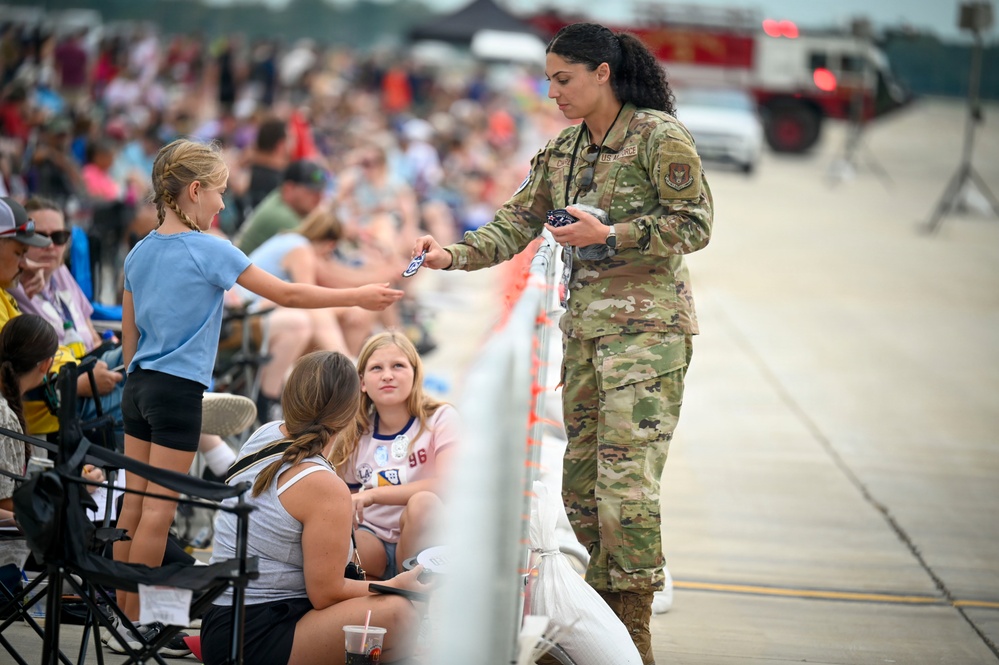 A U.S. Air Force service member in OCP (Operational Camouflage Pattern) uniform interacts with civilian attendees at what appears to be an air show event. The airman is handing a small item, likely a patch or coin, to a young girl across a barrier fence. A large crowd of spectators is visible in the background along a flight line.