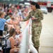 A U.S. Air Force service member in OCP (Operational Camouflage Pattern) uniform interacts with civilian attendees at what appears to be an air show event. The airman is handing a small item, likely a patch or coin, to a young girl across a barrier fence. A large crowd of spectators is visible in the background along a flight line.