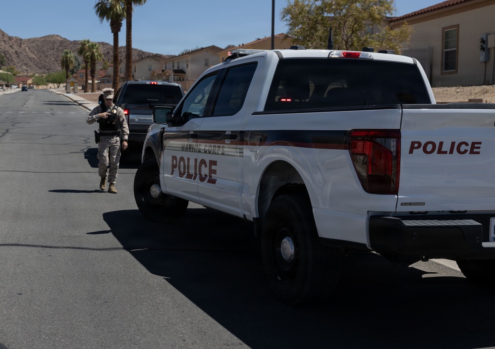 A Marine Corps Police pickup truck is parked on a road within what appears to be a military installation, with a uniformed service member walking alongside it. The setting features desert terrain with palm trees, rocky hills, and tan/beige military-style buildings in the background. The scene suggests a routine patrol or traffic stop activity on base.