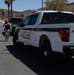 A Marine Corps Police pickup truck is parked on a road within what appears to be a military installation, with a uniformed service member walking alongside it. The setting features desert terrain with palm trees, rocky hills, and tan/beige military-style buildings in the background. The scene suggests a routine patrol or traffic stop activity on base.