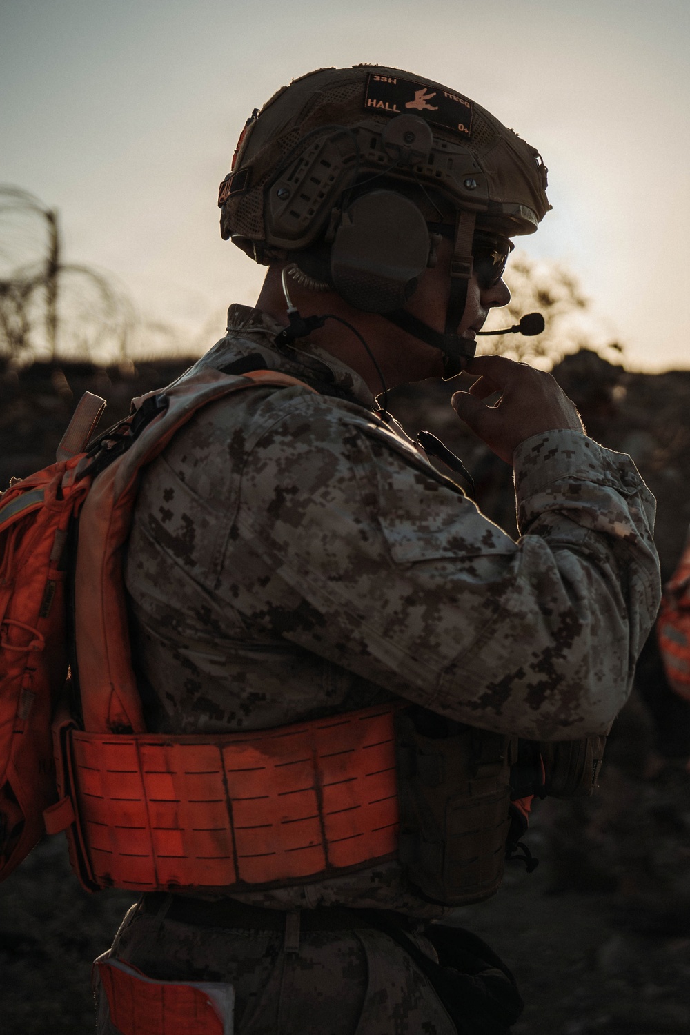 A military service member is photographed from behind at dusk or dawn, wearing full tactical gear including a combat helmet with name tape reading 'HALL' and designation '33H'. The individual is using a boom microphone headset and appears to be communicating via radio. The orange/red plate carrier vest and backpack are distinctive high-visibility elements against the desert camouflage uniform.