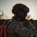 A military service member is photographed from behind at dusk or dawn, wearing full tactical gear including a combat helmet with name tape reading 'HALL' and designation '33H'. The individual is using a boom microphone headset and appears to be communicating via radio. The orange/red plate carrier vest and backpack are distinctive high-visibility elements against the desert camouflage uniform.