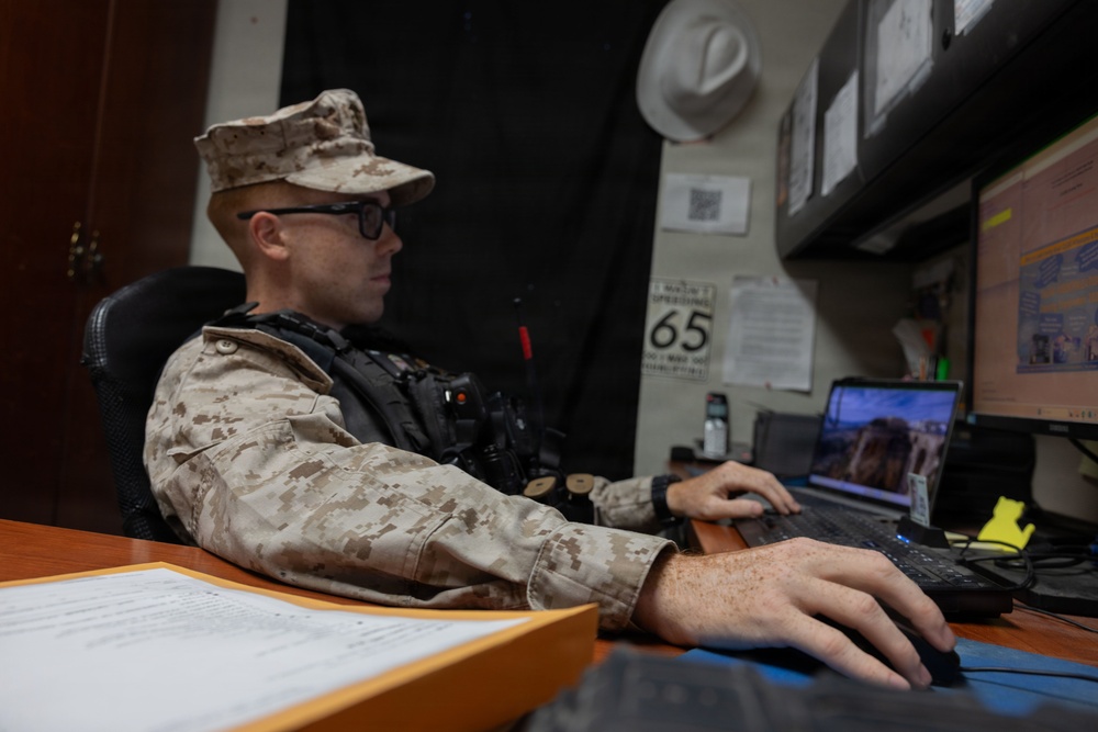 A U.S. Marine in desert MARPAT camouflage uniform and patrol cap is seated at a desk working on a computer. The individual is wearing glasses and a tactical vest with a radio attached. The setting appears to be an administrative or operations office environment.