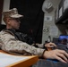 A U.S. Marine in desert MARPAT camouflage uniform and patrol cap is seated at a desk working on a computer. The individual is wearing glasses and a tactical vest with a radio attached. The setting appears to be an administrative or operations office environment.