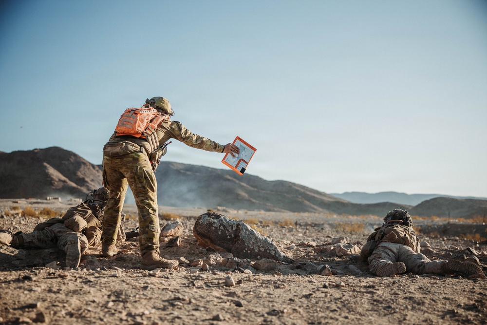 The image depicts a military field exercise or training operation in an arid, desert terrain with rocky hills in the background. A standing soldier wearing multicam camouflage and a distinctive orange backpack is holding out what appears to be a map or tablet in an orange case, likely coordinating with prone soldiers on the ground. The scene suggests a tactical planning or briefing moment during a live-fire or field training exercise.