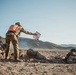 The image depicts a military field exercise or training operation in an arid, desert terrain with rocky hills in the background. A standing soldier wearing multicam camouflage and a distinctive orange backpack is holding out what appears to be a map or tablet in an orange case, likely coordinating with prone soldiers on the ground. The scene suggests a tactical planning or briefing moment during a live-fire or field training exercise.