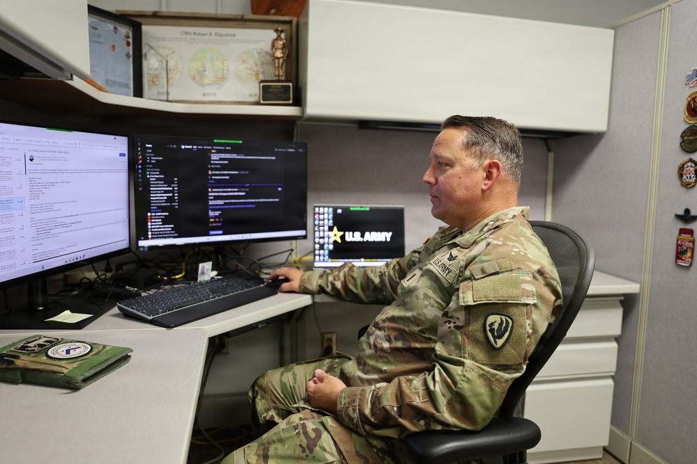 A U.S. Army soldier in OCP (Operational Camouflage Pattern) uniform is seated at a multi-monitor workstation in a military office cubicle. The soldier appears to be working on a computer, with multiple screens displaying what appears to be email, code or terminal windows, and a U.S. Army screensaver. The workspace is personalized with military patches, plaques, and a nameplate visible in the background.