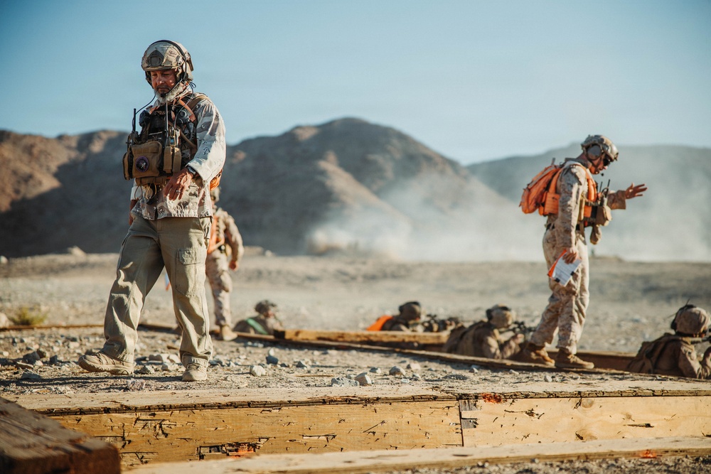 Military personnel are conducting what appears to be a training exercise in an arid desert environment. Multiple soldiers in camouflage uniforms and helmets are visible, with some prone on the ground and others standing and observing. The scene includes dust clouds in the background suggesting recent movement or activity.