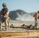 Military personnel are conducting what appears to be a training exercise in an arid desert environment. Multiple soldiers in camouflage uniforms and helmets are visible, with some prone on the ground and others standing and observing. The scene includes dust clouds in the background suggesting recent movement or activity.