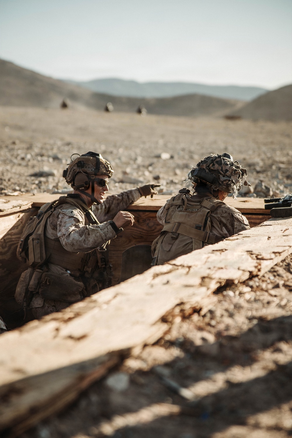 Two military personnel in desert camouflage uniforms are positioned in a fighting trench or fortified position in an arid, rocky desert environment. One soldier is pointing toward a distant target or position while appearing to communicate with the other. The background reveals a barren desert landscape with rolling hills and what appear to be additional personnel or vehicles in the distance.