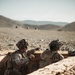 Two military personnel in desert camouflage uniforms are positioned in a fighting trench or fortified position in an arid, rocky desert environment. One soldier is pointing toward a distant target or position while appearing to communicate with the other. The background reveals a barren desert landscape with rolling hills and what appear to be additional personnel or vehicles in the distance.