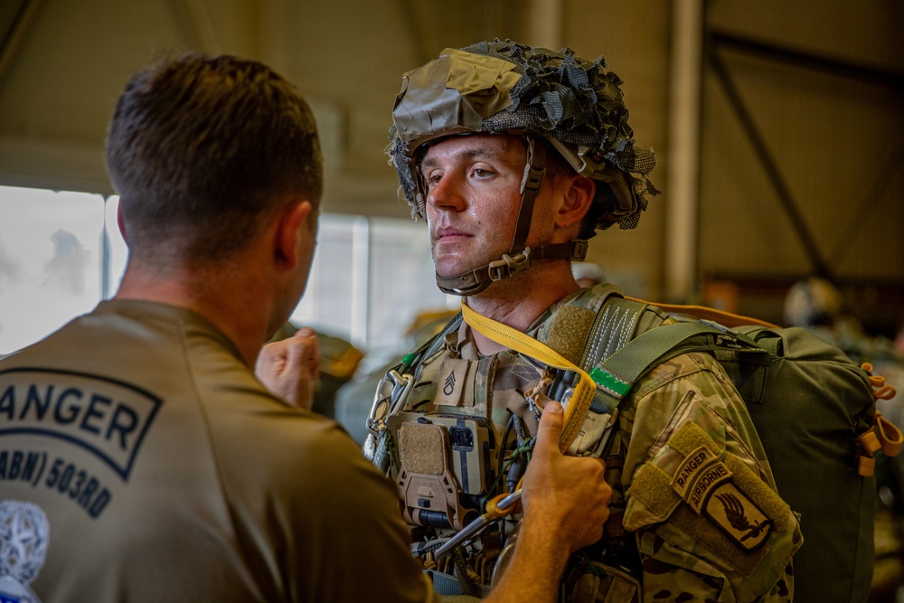Two U.S. Army soldiers are shown in what appears to be a hangar or staging area, with one soldier conducting an equipment check on the other. The soldier facing the camera is wearing full airborne jump gear including a combat helmet with camouflage netting, tactical vest, and parachute harness with a visible yellow static line. Both soldiers are affiliated with the 503rd Infantry Regiment (Airborne) and display Ranger and Airborne tabs.