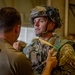 Two U.S. Army soldiers are shown in what appears to be a hangar or staging area, with one soldier conducting an equipment check on the other. The soldier facing the camera is wearing full airborne jump gear including a combat helmet with camouflage netting, tactical vest, and parachute harness with a visible yellow static line. Both soldiers are affiliated with the 503rd Infantry Regiment (Airborne) and display Ranger and Airborne tabs.