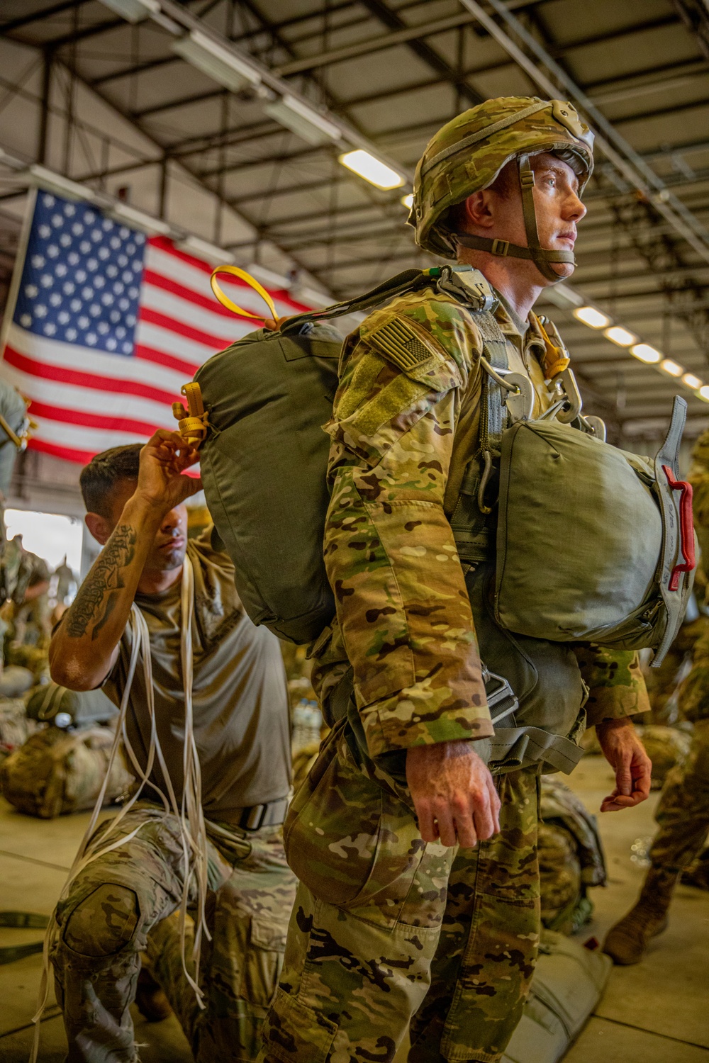 A U.S. Army paratrooper in full jump gear, including a parachute harness and helmet, stands in a hangar while another soldier assists with equipment checks behind him. An American flag is prominently displayed in the background. The scene depicts pre-jump preparations consistent with airborne operations.