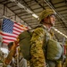 A U.S. Army paratrooper in full jump gear, including a parachute harness and helmet, stands in a hangar while another soldier assists with equipment checks behind him. An American flag is prominently displayed in the background. The scene depicts pre-jump preparations consistent with airborne operations.