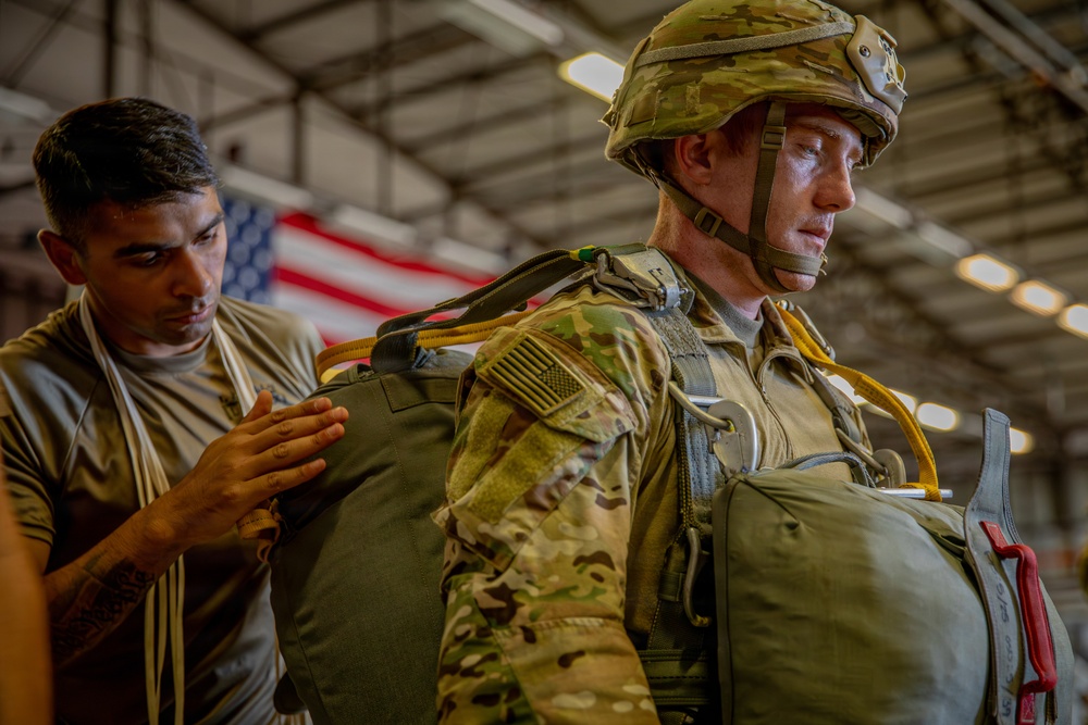 Two military personnel are inside what appears to be a hangar facility, with one soldier assisting another in checking or adjusting parachute equipment. The soldier being assisted is wearing full OCP (Operational Camouflage Pattern) uniform, a combat helmet with chin strap secured, and is equipped with a parachute rig including visible yellow static line. An American flag is visible in the background.