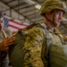 Two military personnel are inside what appears to be a hangar facility, with one soldier assisting another in checking or adjusting parachute equipment. The soldier being assisted is wearing full OCP (Operational Camouflage Pattern) uniform, a combat helmet with chin strap secured, and is equipped with a parachute rig including visible yellow static line. An American flag is visible in the background.