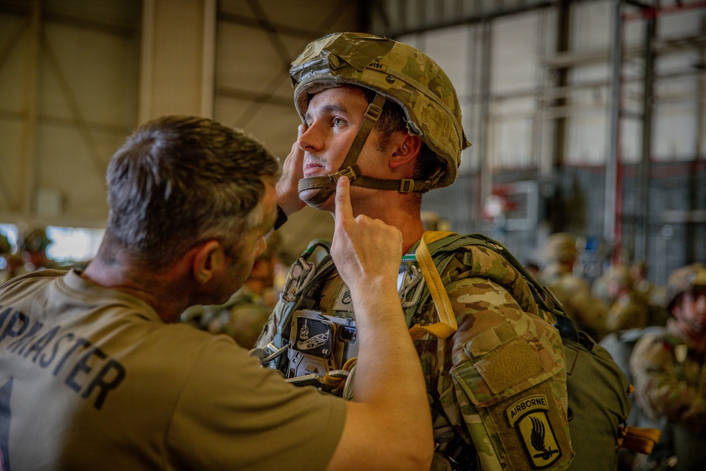 A military jumpmaster is conducting a pre-jump equipment inspection on a soldier wearing full airborne gear inside what appears to be a hangar or staging area. The jumpmaster is checking the soldier's helmet chin strap and overall equipment fit. The soldier is wearing a patch identifying him as belonging to the 173rd Airborne Brigade.