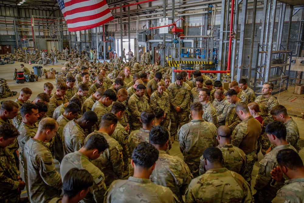 A large group of U.S. Army soldiers in MultiCam/OCP camouflage uniforms are gathered in a large industrial hangar or warehouse facility. The soldiers appear to be bowing their heads in a collective prayer or moment of reflection, gathered in a circular formation. An American flag is prominently displayed hanging from the ceiling above the assembly.