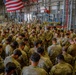 A large group of U.S. Army soldiers in MultiCam/OCP camouflage uniforms are gathered in a large industrial hangar or warehouse facility. The soldiers appear to be bowing their heads in a collective prayer or moment of reflection, gathered in a circular formation. An American flag is prominently displayed hanging from the ceiling above the assembly.