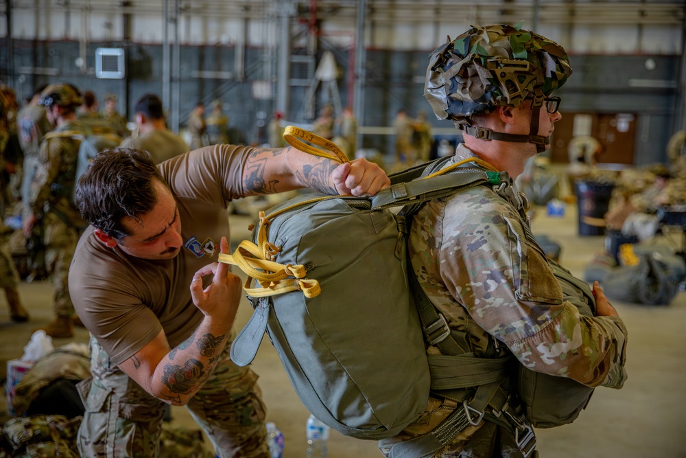 A military personnel in a brown t-shirt with visible tattoos is inspecting or adjusting a parachute pack on another soldier wearing full combat gear including a camouflaged helmet and OCP (Operational Camouflage Pattern) uniform. The setting appears to be a military hangar with numerous other soldiers visible in the background preparing equipment. The yellow straps visible are consistent with parachute rigging hardware.