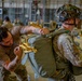A military personnel in a brown t-shirt with visible tattoos is inspecting or adjusting a parachute pack on another soldier wearing full combat gear including a camouflaged helmet and OCP (Operational Camouflage Pattern) uniform. The setting appears to be a military hangar with numerous other soldiers visible in the background preparing equipment. The yellow straps visible are consistent with parachute rigging hardware.