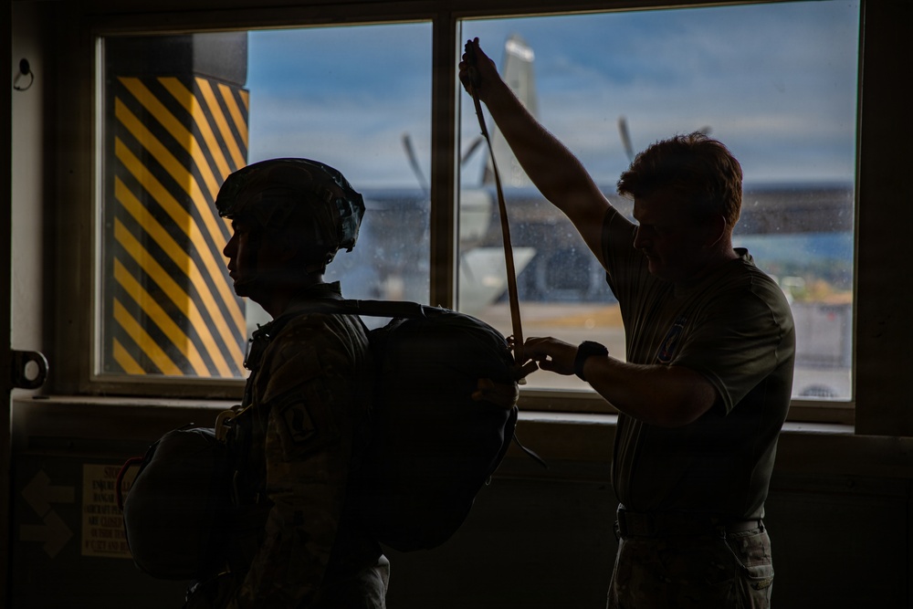 Two individuals are silhouetted against large windows in what appears to be a military or aviation facility. One person is wearing a tactical helmet and carrying a large pack, while the second person appears to be adjusting or checking a strap or harness. An aircraft and yellow-black hazard-striped pillar are visible through the windows in the background.