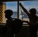 Two individuals are silhouetted against large windows in what appears to be a military or aviation facility. One person is wearing a tactical helmet and carrying a large pack, while the second person appears to be adjusting or checking a strap or harness. An aircraft and yellow-black hazard-striped pillar are visible through the windows in the background.