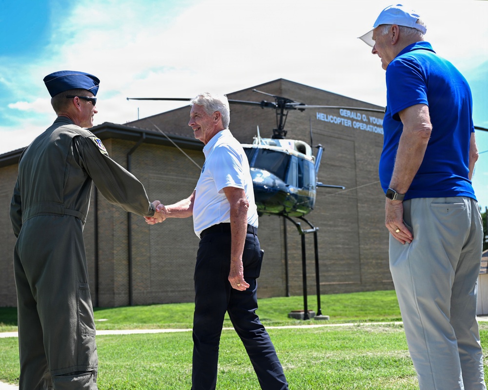 Three men are gathered outdoors in front of a building identified as the 'Gerald O. Young Helicopter Operations' facility. A military officer in a green flight suit is shaking hands with a civilian in a white shirt, while a third man in a blue polo shirt observes. A vintage helicopter is displayed on a static mount in front of the building.