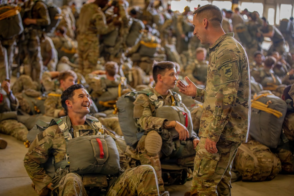 A large group of military personnel in camouflage uniforms are gathered inside what appears to be a hangar or staging area, equipped with parachute rigs and jump gear. A standing soldier is engaged in conversation with seated soldiers, one of whom is smiling and laughing. The scene suggests a pre-jump briefing or waiting period before an airborne operation.