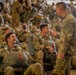 A large group of military personnel in camouflage uniforms are gathered inside what appears to be a hangar or staging area, equipped with parachute rigs and jump gear. A standing soldier is engaged in conversation with seated soldiers, one of whom is smiling and laughing. The scene suggests a pre-jump briefing or waiting period before an airborne operation.