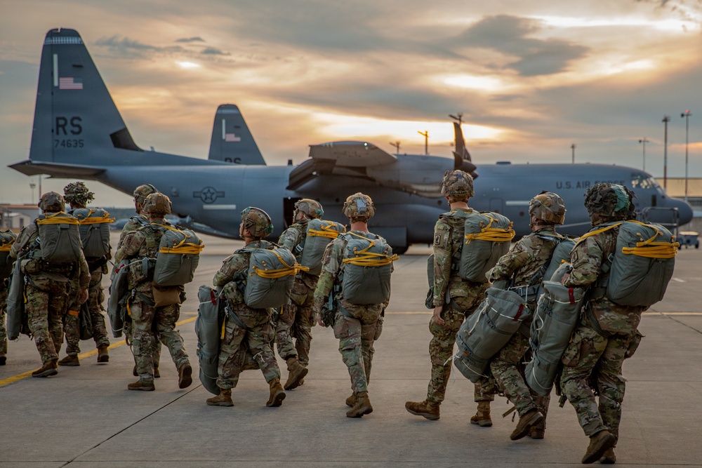 A group of military paratroopers in full combat gear and parachute equipment are walking across a tarmac toward a U.S. Air Force C-130 Hercules transport aircraft. The soldiers are carrying large rucksacks and parachute packs with yellow static lines visible. The scene is captured at dusk or dawn with a dramatic sky in the background.