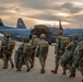 A group of military paratroopers in full combat gear and parachute equipment are walking across a tarmac toward a U.S. Air Force C-130 Hercules transport aircraft. The soldiers are carrying large rucksacks and parachute packs with yellow static lines visible. The scene is captured at dusk or dawn with a dramatic sky in the background.