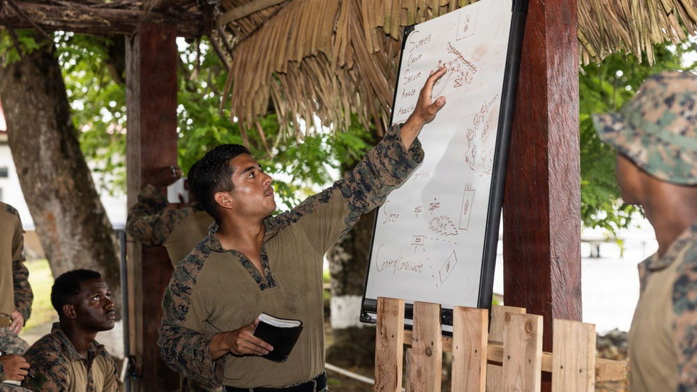 A military service member in camouflage uniform is conducting a briefing or training session under an open-air thatched-roof structure, pointing at a whiteboard with written notes and diagrams. Several other uniformed personnel are present as an audience, suggesting a tactical or instructional exercise. The outdoor setting with tropical vegetation indicates a warm-climate training environment.