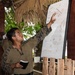 A military service member in camouflage uniform is conducting a briefing or training session under an open-air thatched-roof structure, pointing at a whiteboard with written notes and diagrams. Several other uniformed personnel are present as an audience, suggesting a tactical or instructional exercise. The outdoor setting with tropical vegetation indicates a warm-climate training environment.