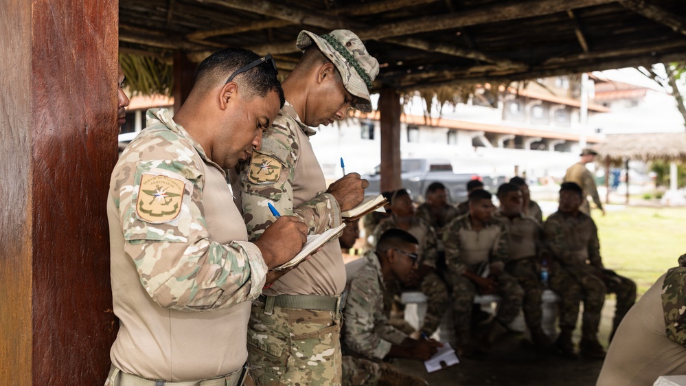 A group of military personnel in camouflage uniforms are gathered under an open-air thatched shelter, likely during a briefing or training exercise. Two soldiers in the foreground are actively writing notes on small notepads using blue pens, while additional soldiers are seated in the background. The setting appears to be an outdoor military or civic facility in a tropical environment.
