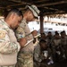 A group of military personnel in camouflage uniforms are gathered under an open-air thatched shelter, likely during a briefing or training exercise. Two soldiers in the foreground are actively writing notes on small notepads using blue pens, while additional soldiers are seated in the background. The setting appears to be an outdoor military or civic facility in a tropical environment.